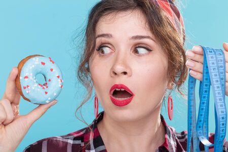 Close-up of a shocked and upset young beautiful woman holding a blue donut in her hands and measuring tape. Concept of rejection of high-calorie foods and healthy lifestyle choice.の写真素材