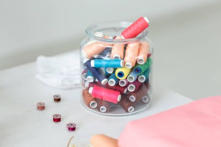 Close up multicolored threads and bobbins lie on a table in a transparent glass jar on the...