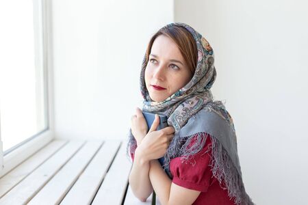 Close-up portrait of a cute young dreamy woman with a book in her hand looking out of the window and thinking about something. Concept lovers of classics and literature.の写真素材
