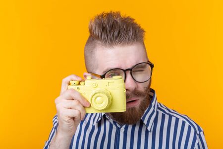 Cute young male photographer with a mustache and beard is photographing a yellow vintage camera on a yellow background. Concept of hobby and professional work.の写真素材