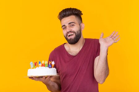 Positive handsome young hipster man in burgundy t-shirt holding congratulatory cake with inscription...