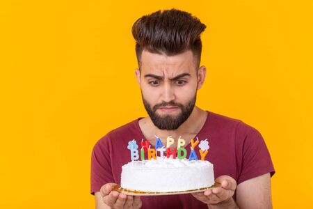 Embarrassed young male hipster with beard holding a birthday cake in his hands and looking...