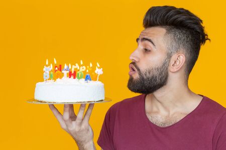 Side view of a handsome arab young bearded man blowing off candles with a congratulatory cake posing on a yellow backgroundの写真素材