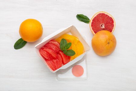 Top view slices of grapefruit and orange are in a lunch box on a table next to half a grapefruit and two oranges. Fruit snack at work concept.の写真素材