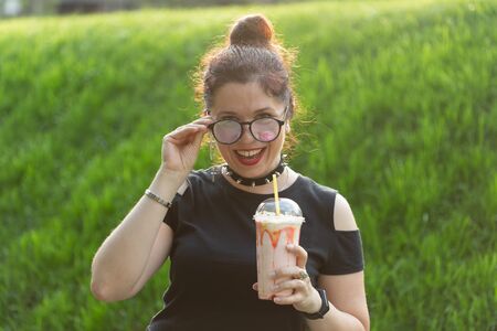 Portrait of a positive young pretty girl in punk clothes and glasses with a milkshake in her hands...
