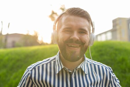 Portrait of a positive young man with a mustache and beard listening to an audiobook while walking...