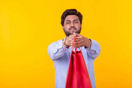 Indian man with a beard is holding red shopping bags posing on a yellow background. Supermarket...