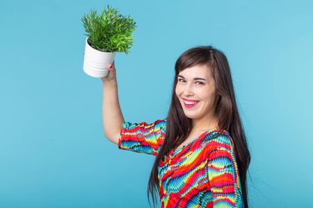 Cheerful young woman is holding a pot with a plant posing against a blue background. Concept of gardening and interior accessories.の写真素材
