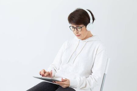 Woman 40-50 years old in headphone sitting on chair and hold tablet on white background with copy spaceの写真素材