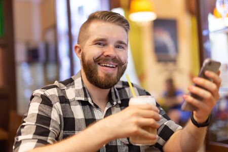 Stylish young man with a mustache and beard is watching a social network using a smartphone while sitting in a cafe on a weekend. The concept of dependence on social networks.の写真素材
