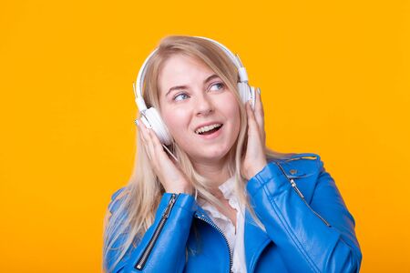 Portrait of pretty young girl blonde female student holding smartphone with blue leather jacket headphones posing on a yellow background. Concept of listening to online radio and music subscription.の写真素材