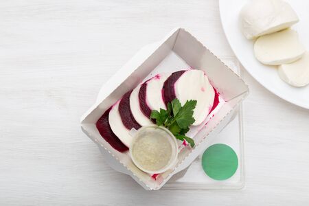 Top view of boiled beets with slices of white cheese lie in a white lunch box with sour cream sauce and parsley on a white table next to goat cheese. Protein snack conceptの写真素材