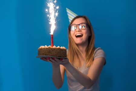 Cheerful young blurred girl student in glasses holding a congratulatory cake with a candle standing on a blue background. Birthday concept.の写真素材