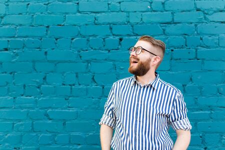 Portrait of a young positive stylish smiling guy with a mustache and beard against the background of a blue brick wall with copy space. Vacation concept in the city.の写真素材