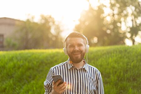 Positive young male hipster listens to your favorite online radio using smartphone headphones and high-speed wireless internet in the park on a sunny warm summer day.の写真素材