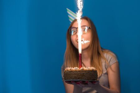 Cheerful young blurred girl student in glasses holding a congratulatory cake with a candle standing on a blue background. Birthday concept.の写真素材