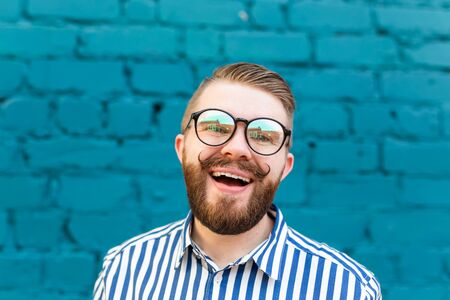 Close-up portrait of a happy young man in glasses with a mustache and beard posing on a background of blue blurred brick wall. Concept of happinessの写真素材