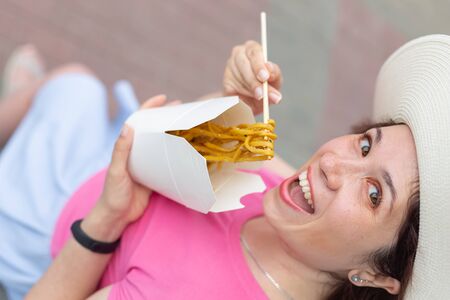 Close-up of young woman in a hat eats Chinese noodles while walking in the park on a warm summer day. Takeaway food concept.の写真素材