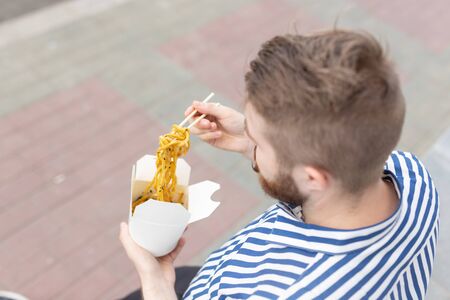 Close-up of an amusing young hipster guy eating chinese noodles with wooden chopsticks sitting in a park outside on a warm summer day. The concept of rest and snacking on the street.の写真素材