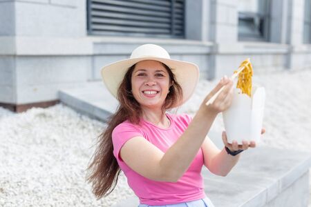Young positive girl in a hat eats chinese noodles with wooden chopsticks for take-away lunch. Concept of high-calorie healthy food.の写真素材