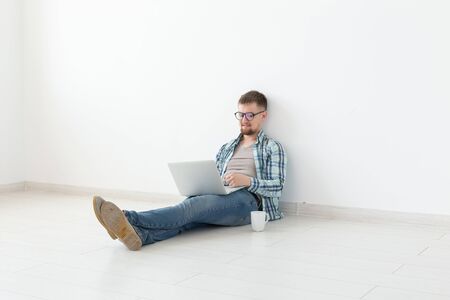 Positive young man in casual clothes surfing the Internet in search of new housing sitting on the floor in an empty room. The concept of finding an apartment using the Internet and a laptop.の写真素材