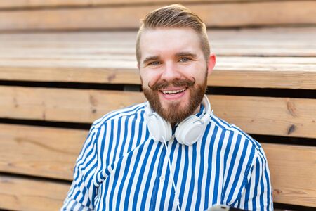 Portrait of a handsome young student guy with a mustache and a beard relaxing in a park with headphones on a warm summer day. Weekend getaway concept.の写真素材