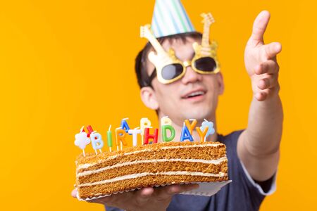 Portrait of a funny positive guy with a paper cap and glasses holding a congratulatory homemade cake...