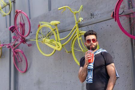 Portrait of a positive cheerful young arab man with a glass of juice with a straw while walking in the city on a warm sunny summer day. The concept of rest after study and work on weekends.の写真素材