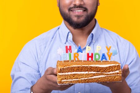 Close-up of funny young indian guy with a cap and a homemade cake in his hands posing on a yellow background. Anniversary and birthday concept.の写真素材