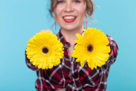 Portrait of positive young woman in plaid shirt holding two yellow flowers with different hearts near breast posing against blue background. Concept of body beauty in all forms, Body positivityの写真素材