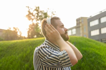 Handsome relaxed young hipster man with a mustache and beard is listening to favorite music at the earphones while walking through the park. Music subscription concept and online radio.の写真素材