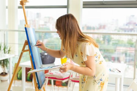 Art school, creativity and leisure concept - student girl or young woman artist with easel, palette and paint brush painting picture at studioの写真素材