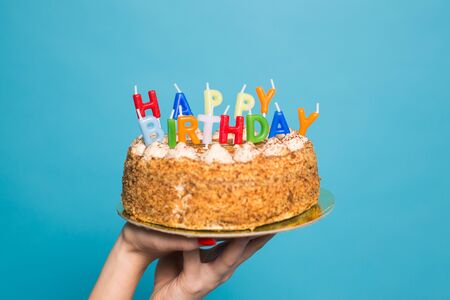 Hands holding a birthday cake with candles and the inscription birthday on a blue background. Congratulations on the anniversary and birthday. Copyspaceの写真素材