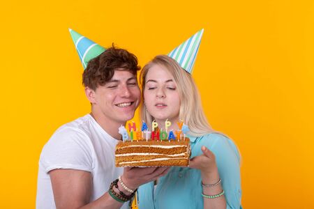 Cheerful young couple charming guy and cute girl in paper hats make foolish face and hold in their hands a cake with the inscription birthday standing on a yellow background. Concept greetings and prank.の写真素材