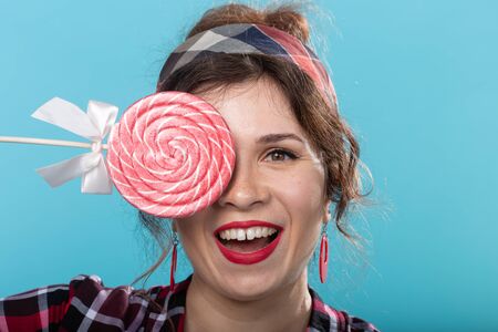 Pretty smiling young woman in a plaid shirt closes her eye with big colorful lollipops and posing...