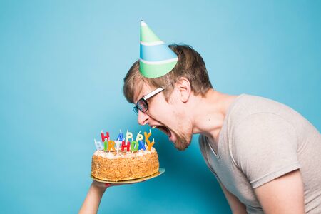 Funny crazy young man in a greeting paper hat wants to bite off a piece of congratulatory cake. Concept of fooling around and birthday greetings. Copy spaceの写真素材