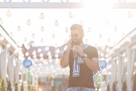 Portrait of a positive cheerful young arab man with a glass of juice with a straw while walking in the park on a warm sunny summer day. The concept of rest after study and work on weekends.の写真素材