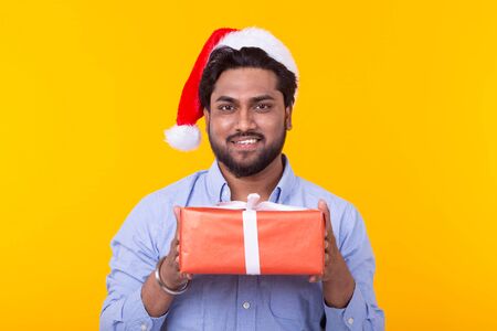Positive young indian man Santa Claus with gifts and Christmas tree posing on a yellow background. Christmas and New Year holidays concept.の写真素材
