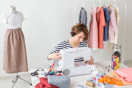 Young seamstress woman sews a new product on a sewing machine sitting at the table with its accessories. Concept of designer clothes.の写真素材