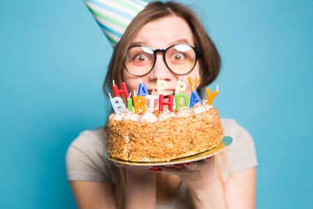 Charming merry crazy young girl student in congratulatory paper hat holding a happy birthday cake in her hands standing against a blue background. Advertising space.の写真素材