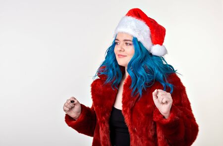 Christmas, fashion and youth - Beautiful young woman with blue hair in red fur coat and santa hat, looking at the camera and standing against a white background. Ready for party.の写真素材