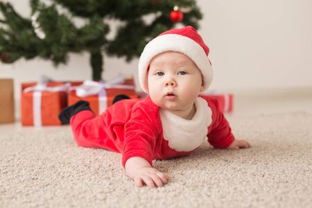 Cute baby girl wearing santa claus suit crawling on floor over Christmas tree. Holiday season.