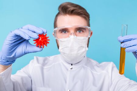 Covid-19, Vaccine development and medicine concept - Man scientist in flu mask and protective gloves holding a model of coronavirus and test tube.の写真素材