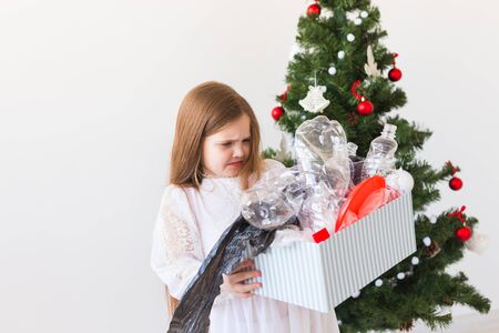 Shocked little child girl looks with opened eyes and worried expression, holding box with various plastic wastes over christmas tree background.の写真素材