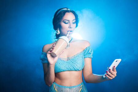 Magic, cosplay and fairy tale concept - Portrait of a young woman in the image of an Eastern fairy Princess holds smartphone and shaker on blue background.の写真素材