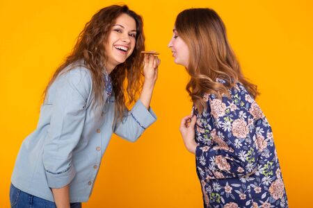 Two funny female friends or sisters eating sharing cookie on yellow background. Friendship, relationship and fun concept.の写真素材