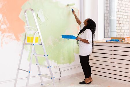 Beautiful african american girl painting the wall with paint roller. Portrait of a young beautiful woman painting wall in her new apartment. Redecoration and renovation concept.の写真素材