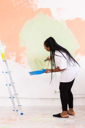 Beautiful african american girl painting the wall with paint roller. Portrait of a young beautiful woman painting wall in her new apartment. Redecoration and renovation concept.の写真素材