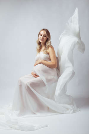 Elegant pregnant young woman standing wearing flying white fabric. Pregnancy, maternity and motherhood concept.の写真素材