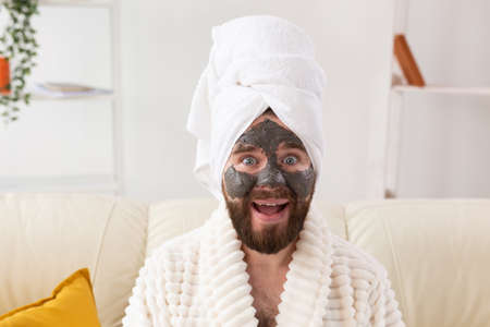 Bearded man having fun with a cosmetic mask on his face made from black clay. Men skin care, humor and spa at home conceptの写真素材